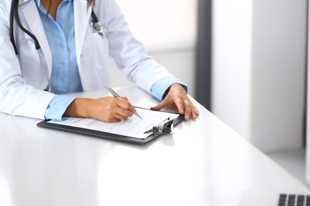 Unknown doctor woman filling up medical form while sitting at the desk in hospital office, close-up of hands. Physician at work. Medicine and health care conceptの写真素材