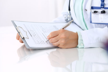 Unknown female doctor filling up an application form with silver pen while sitting at the desk in hospital office. Just hands. Medicine and health care concept.の写真素材