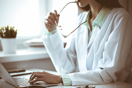 Doctor woman using laptop computer while sitting at the desk near window in hospital. Medicine and health care concept. Green is main colorの写真素材