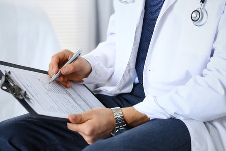 Doctor man filling up medical form on clipboard at hospital, close-up. Healthcare, insurance and medicine conceptの写真素材