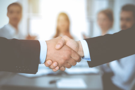 Business people shaking hands at meeting while theirs colleagues clapping and applausing. Group of unknown businessmen and women in modern white office. Success teamwork, partnership and handshake concept.の写真素材