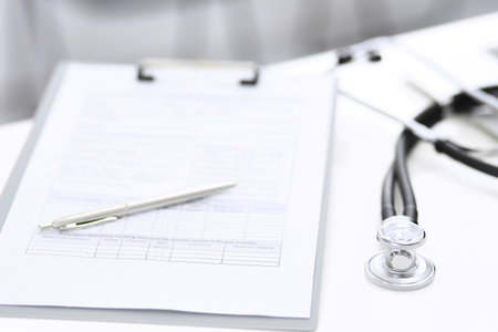 Stethoscope, clipboard with medical form lying on hospital reception desk with laptop computer and busy doctor and patient communicating at the background. Medical tools at doctor working table.Medicine conceptの写真素材