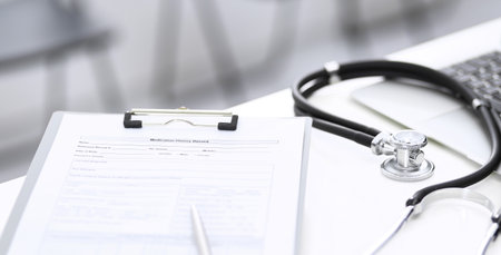 Stethoscope, clipboard with medical form lying on hospital reception desk with laptop computer and busy doctor and patient communicating at the background. Medical tools at doctor working table.Medicine conceptの写真素材