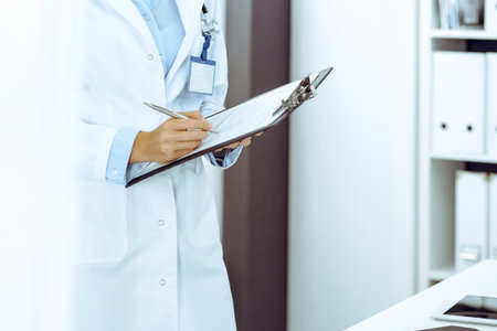 Doctor woman filling up medical form in clinic office. Unknown female physician at work while standing straight at hospital, close-up. Medicine and healthcare conceptの写真素材