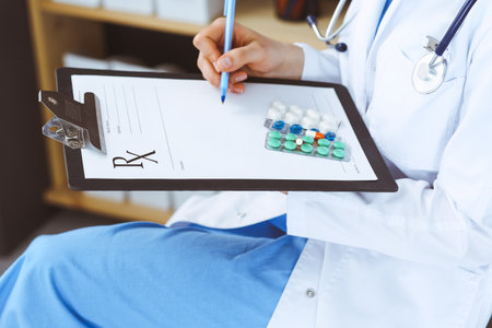 Unknown woman doctor at work at hospital. Pills at medical clipboard. Young female physician write prescription or filling up medical form while sitting in hospital office, close-up.の写真素材