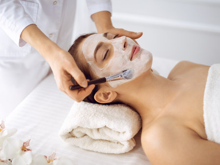 Beautiful brunette woman enjoying applying cosmetic mask with closed eyes. Relaxing treatment in medicine and spa center concepts.の写真素材