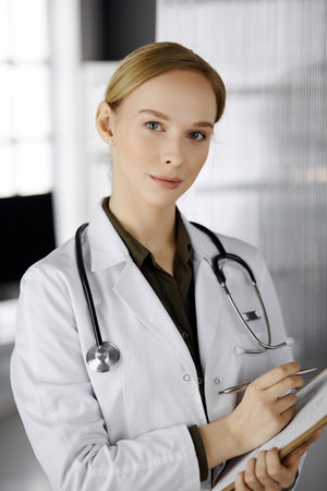 Cheerful smiling female doctor using clipboard in clinic. Portrait of friendly physician woman at work. Perfect medical service in hospital. Medicine conceptの写真素材
