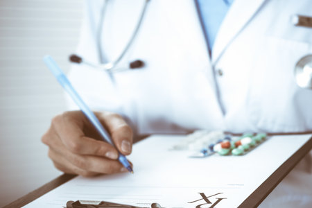 Unknown woman doctor at work at hospital. Pills at medical clipboard. Young female physician write prescription or filling up medical form while sitting in hospital office, close-upの写真素材