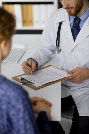 Unknown red-bearded doctor and patient woman discussing current health examination while sitting in sunny clinic, close-up. Medicine concept.の写真素材