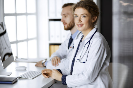 Friendly female doctor sitting in clinic with her male colleague at the background. Perfect medical service and medicine conceptの写真素材