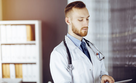 Friendly red-bearded doctor using tablet computer in sunny clinic at his working place. Medicine conceptの写真素材