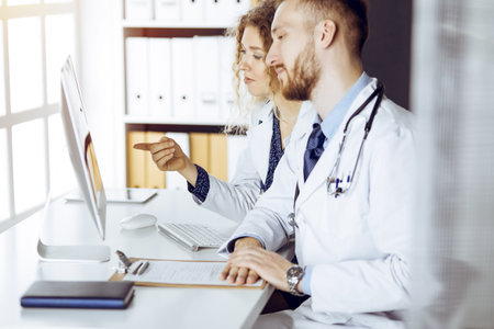 Red-bearded doctor with female colleague discussing current disease therapy while sitting at working place in sunny clinic. Team work in medicineの写真素材