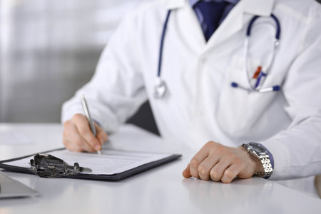 Unknown male doctor sitting and working with clipboard of medication history record in clinic at his working place, close-up. Young physician at work. Perfect medical service, medicine concept.の写真素材