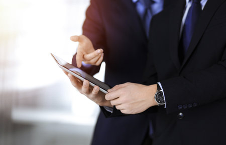 Business people use a tablet computer for discussion of their new project, standing in sunny modern office. Unknown businessman or male entrepreneur with a colleague at workplace.の写真素材