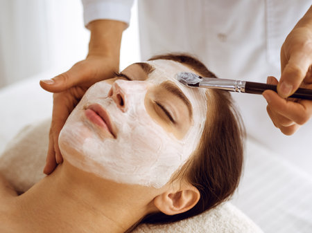 Beautiful brunette woman enjoying applying cosmetic mask with closed eyes. Relaxing treatment in medicine and spa center conceptsの写真素材