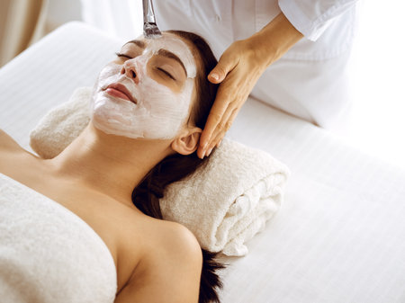 Beautiful brunette woman enjoying applying cosmetic mask with closed eyes. Relaxing treatment in medicine and spa center conceptsの写真素材