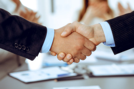 Business people shaking hands at meeting while theirs colleagues clapping and applausing. Group of unknown businessmen and women in modern white office. Success teamwork, partnership and handshake concept.の写真素材