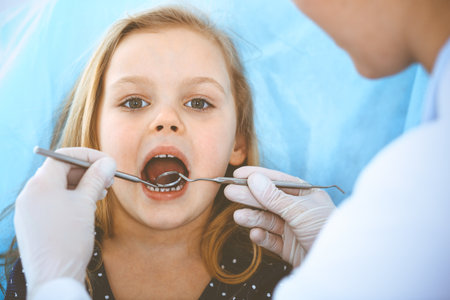 Little baby girl sitting at dental chair with open mouth during oral check up while doctor. Visiting dentist office. Medicine concept. Toned photoの写真素材