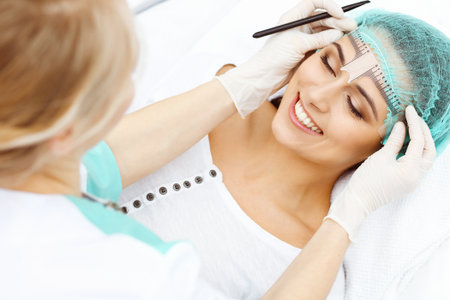 Professional beautician is going to correct eyebrow line at woman face using cosmetic ruler. Permanent brow makeup in beauty salon, closeup. Cosmetology treatmentの写真素材
