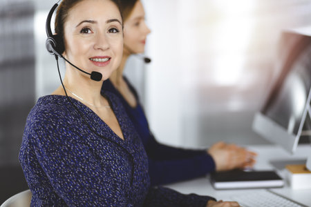 Two busineswomen have conversations with the clients by headsets, while sitting at the desk in a sunny modern office. Diverse people group in a call centerの写真素材