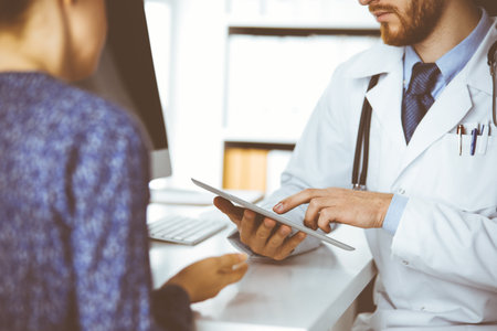 Unknown red-bearded doctor and patient woman discussing current health examination while sitting and using tablet computer in clinic, close-up. Medicine conceptの写真素材