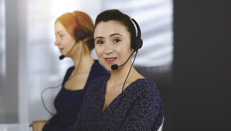 Two busineswomen have conversations with the clients by headsets, while sitting at the desk in a...
