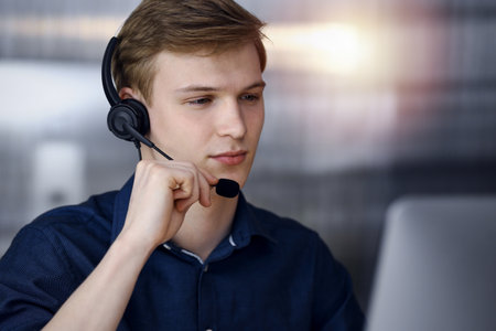 Young blond businessman using headset and computer in a darkened office, glare of light on the background. Startup business means working hardの写真素材