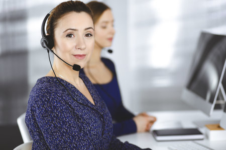 Two busineswomen have conversations with the clients by headsets, while sitting at the desk in a modern office. Diverse people group in a call center. Telemarketing and customer serviceの写真素材
