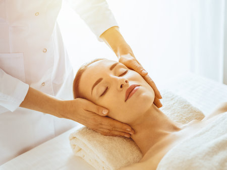 Beautiful woman enjoying facial massage with closed eyes in sunny spa center. Relaxing treatment concept in medicineの写真素材