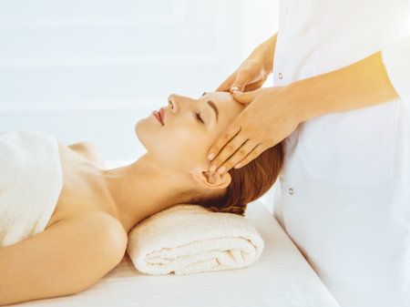 Beautiful woman enjoying facial massage with closed eyes in sunny spa center. Relaxing treatment concept in medicineの写真素材