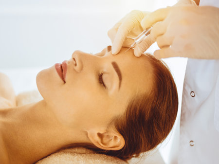Beautiful woman receiving beauty injections with closed eyes in sunny medical center. Beautician doctor hands doing beauty procedure to female face with syringeの写真素材