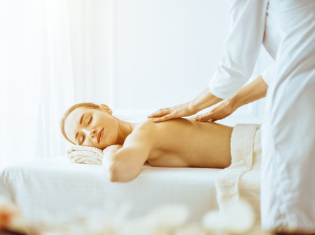 Beautiful woman enjoying back massage with closed eyes in sunny spa centerの写真素材