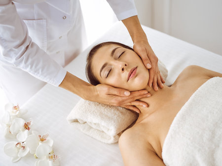 Beautiful brunette woman enjoying facial massage with closed eyes. Relaxing treatment in medicine and spa center conceptsの写真素材
