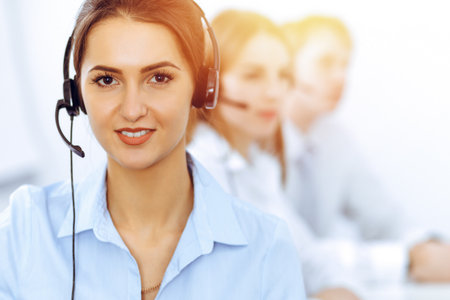 Call center operators. Focus on beautiful business woman using headset in sunny officeの写真素材