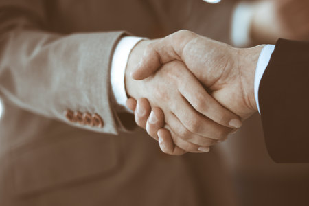 Business people shaking hands while standing with colleagues after meeting or negotiation, close-up. Group of unknown businessmen and women in modern office. Teamwork, partnership and handshake concept, toned picture.の写真素材