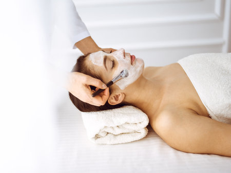 Beautiful brunette woman enjoying applying cosmetic mask with closed eyes. Relaxing treatment in medicine and spa center conceptsの写真素材