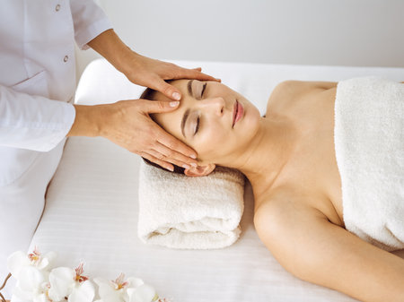 Beautiful brunette woman enjoying facial massage with closed eyes. Relaxing treatment in medicine and spa center conceptsの写真素材