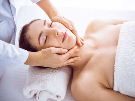 Beautiful brunette woman enjoying facial massage with closed eyes in sunny spa center . Relaxing treatment and cosmetic medicine conceptsの写真素材