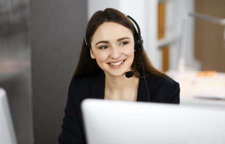 Young friendly girl in headsets is talking to a firms client, while sitting at the desk in a modern...