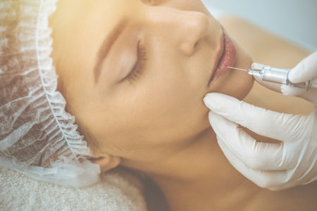 Beautician doing beauty procedure with syringe to face of young brunette woman in sunny clinic. Cosmetic medicine and surgery, beauty injectionsの写真素材