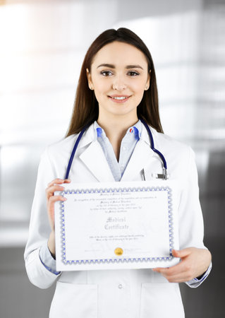 Happy young woman-doctor is holding a certificate in her hands. Professional physician at work in sunny clinicの写真素材
