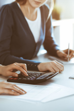 Accountant checking financial statement or counting by calculator income for tax form, hands close-up. Business woman sitting and working with colleague at the desk in office. Audit conceptの写真素材