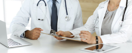 Two unknown doctors discussing treatment problems while sitting at the desk in hospital office,...