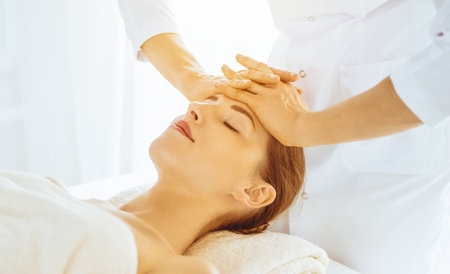 Beautiful woman enjoying facial massage with closed eyes in sunny spa center. Relaxing treatment concept in medicineの写真素材