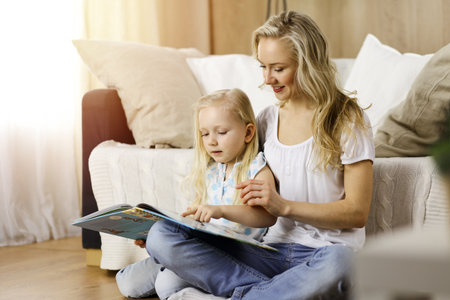 Happy family. Blonde young mother reading a book to her cute daughter while sitting at wooden floor in sunny room. Motherhood conceptの写真素材