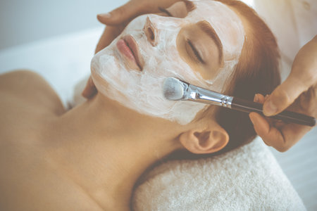 Beautiful brunette woman enjoying applying cosmetic mask with closed eyes. Relaxing treatment in medicine and spa conceptsの写真素材