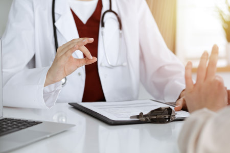 Unknown woman-doctor and patient discussing something while sitting at the table in sunny clinic, close-upの写真素材