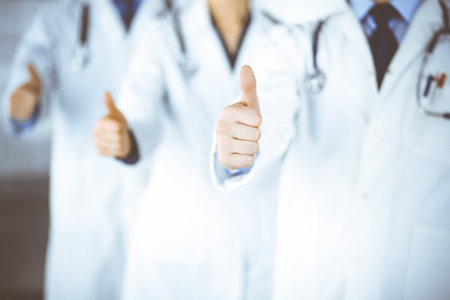 Group of unknown doctors stand as a team with thumbs up in a hospital office. Physicians ready to examine and help patients. Medical help, insurance in health care, best desease treatment and medicine concept.の写真素材