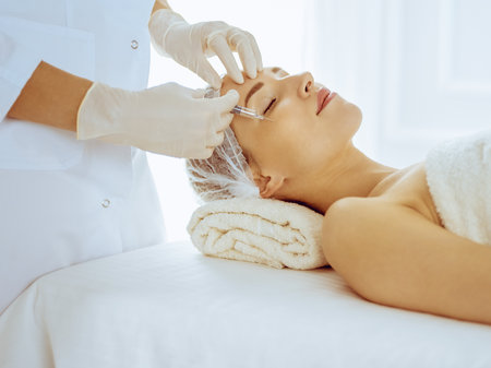 Beautiful woman receiving beauty injections with closed eyes in medical center. Beautician doctor hands doing beauty procedure to female face with syringeの写真素材