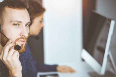 Red-bearded businessman talking by headset near his female colleague while sitting in sunny office....